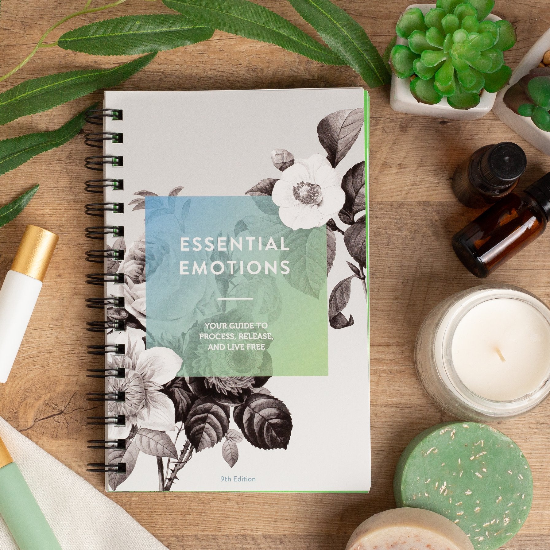 Essential Emotions book laying on a table with essential oil vials, roller bottles, soaps, and plants.